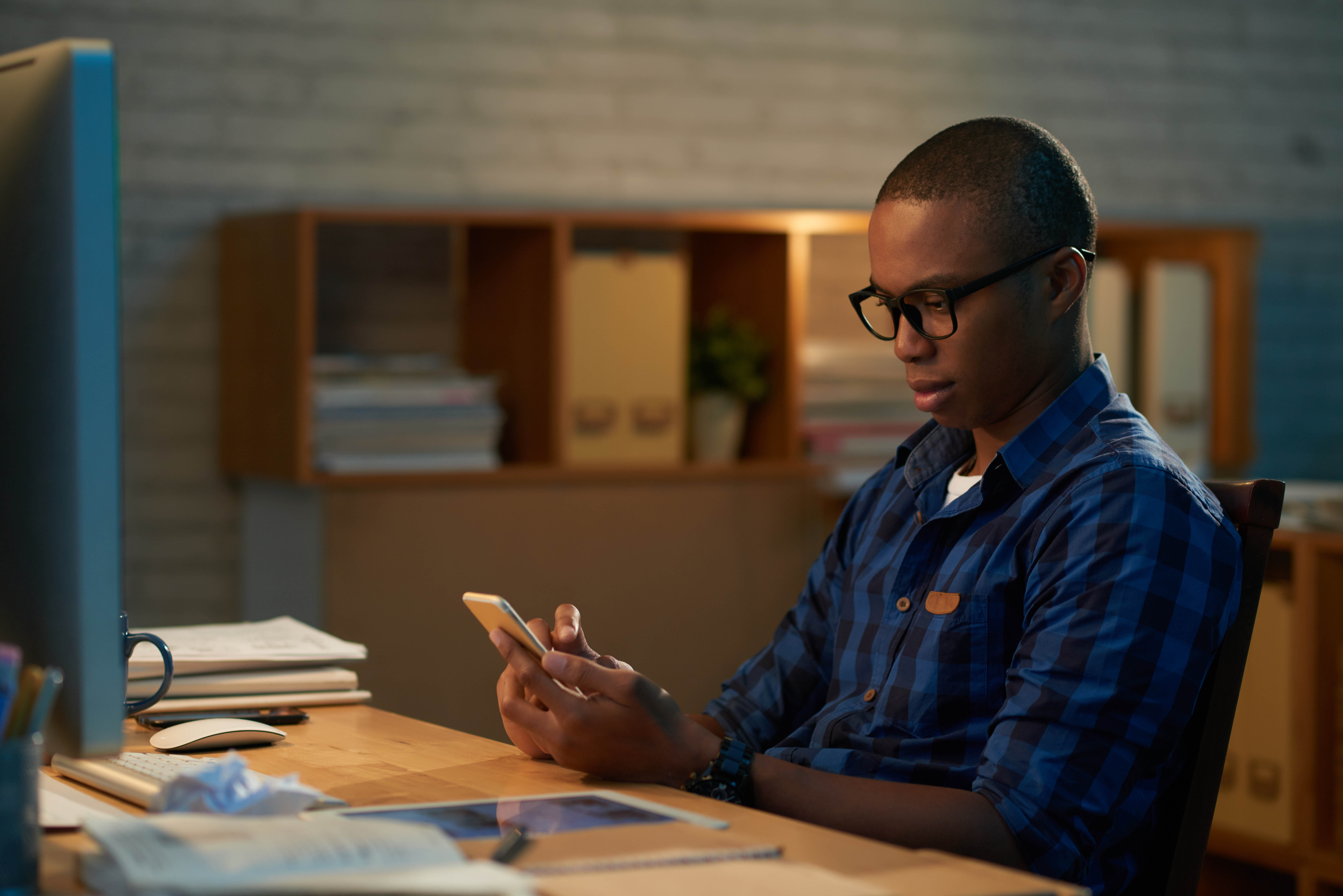 Young Afro-American white collar worker texting with his colleague on smartphone while working overtime in dark open plan office, profile view